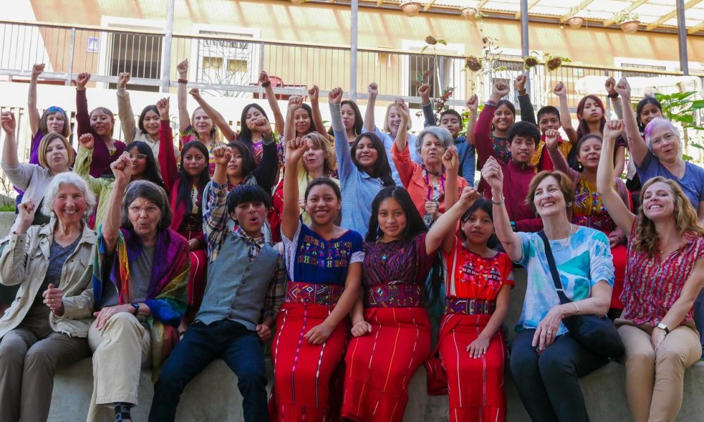 A large group of travelers and school children in Guatemala sit in rows, raising their fists and smiling.